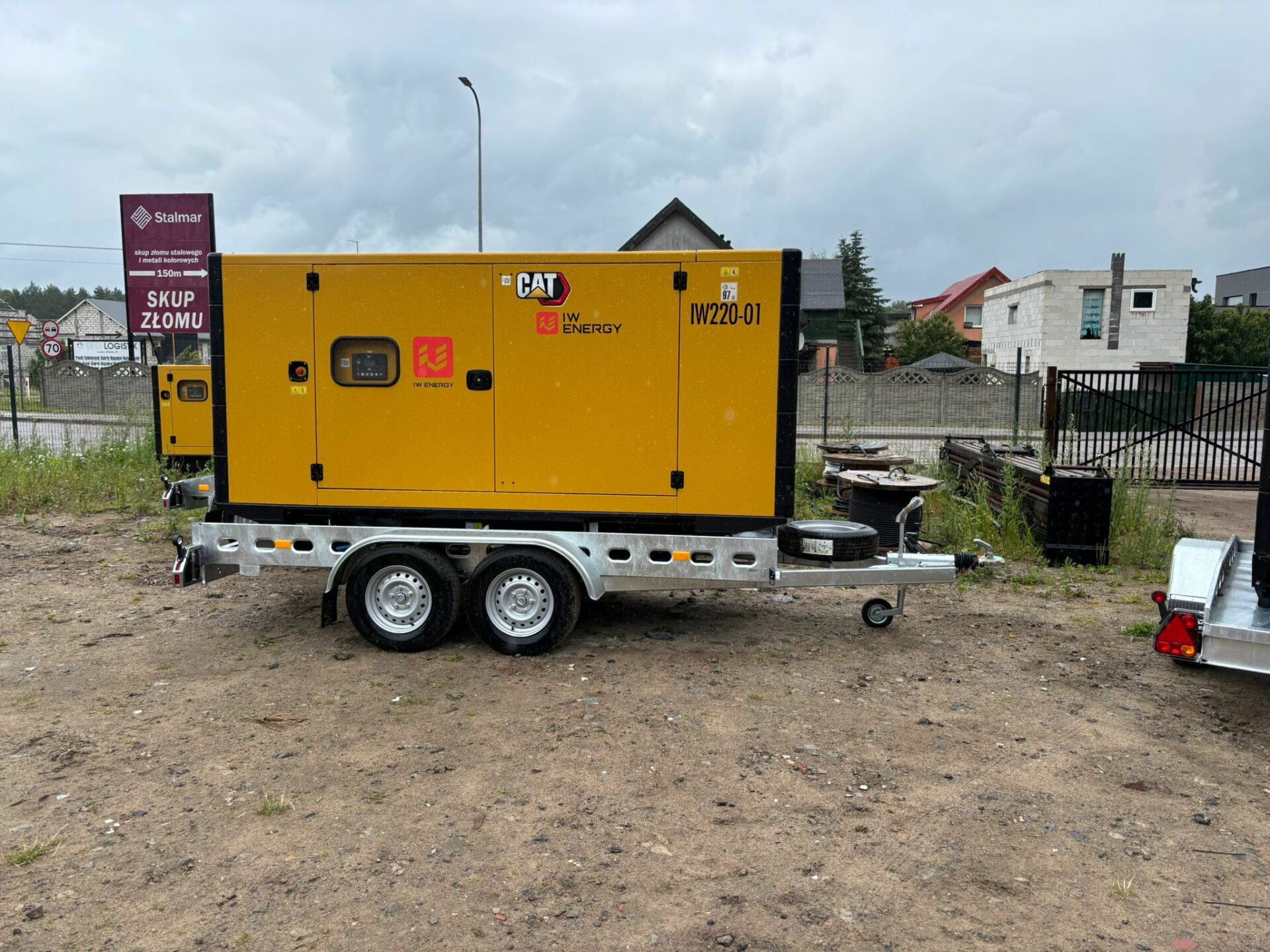 Agregat prądotwórczy marki CAT na przyczepie dwuosiowej, wynajmowany przez firmę IW Energy. Urządzenie oznaczone numerem IW220-01, ustawione na żwirowym placu w pobliżu drogi i zabudowy mieszkalnej. W tle widoczne są budynki, ogrodzenia i szyld skupu złomu.